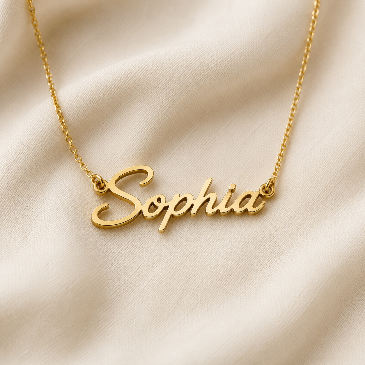 A gold nameplate necklace spelling 'Sophia' in elegant cursive script, resting on a draped cream-colored fabric background.
