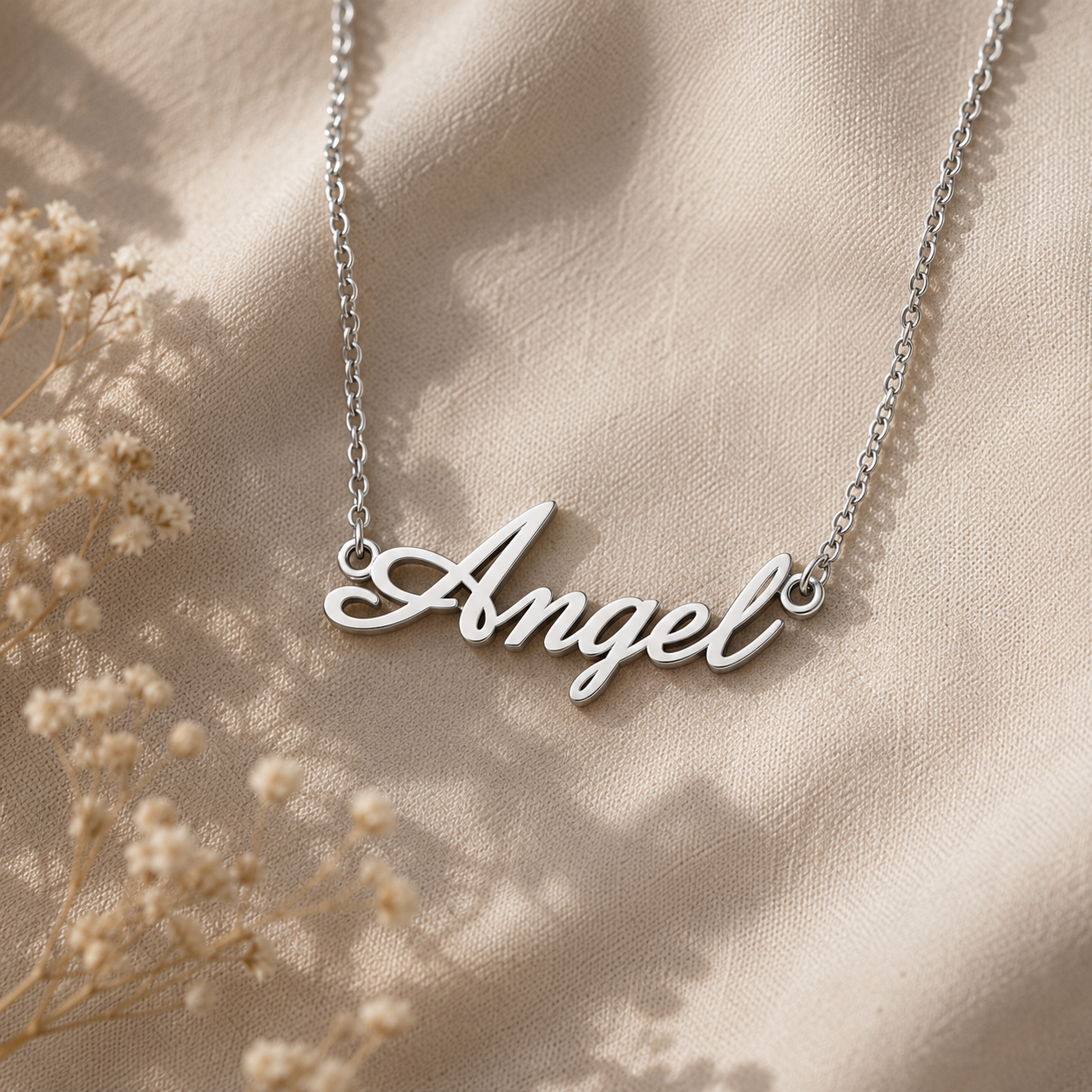 A silver script pendant necklace spelling 'Angel' on a delicate chain, displayed on draped beige fabric with dried floral accents.