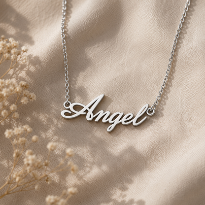 A silver script pendant necklace spelling 'Angel' on a delicate chain, displayed on draped beige fabric with dried floral accents.